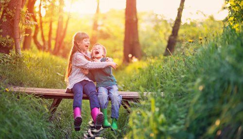 Ein Junge und ein Mädchen sitzen auf einer keinen Holzbrücke und Lachen. Im Hintergrund scheint die Sonne.